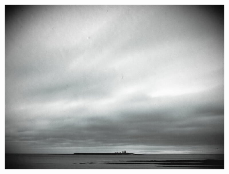 A black-and-white photo of a distant lighthouse on a small island under a cloudy sky, surrounded by calm sea and a dark, moody atmosphere.