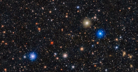 A dense field of stars and galaxies in deep space, featuring various bright blue, white, yellow, and red points of light scattered across a dark background.