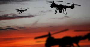 Three drones are silhouetted against a dramatic sunset sky with shades of orange, red, and purple clouds. The drones appear in different positions, with one close to the camera.