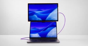 A laptop with a blue-themed screen is on a white surface, connected by a purple cable to a second monitor positioned directly above it, displaying a matching blue background.