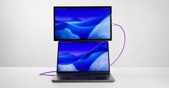 A laptop with a blue-themed screen is on a white surface, connected by a purple cable to a second monitor positioned directly above it, displaying a matching blue background.