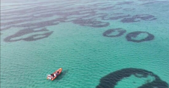 A small red boat with several people floats on clear turquoise water near large, dark circular patterns visible beneath the surface.
