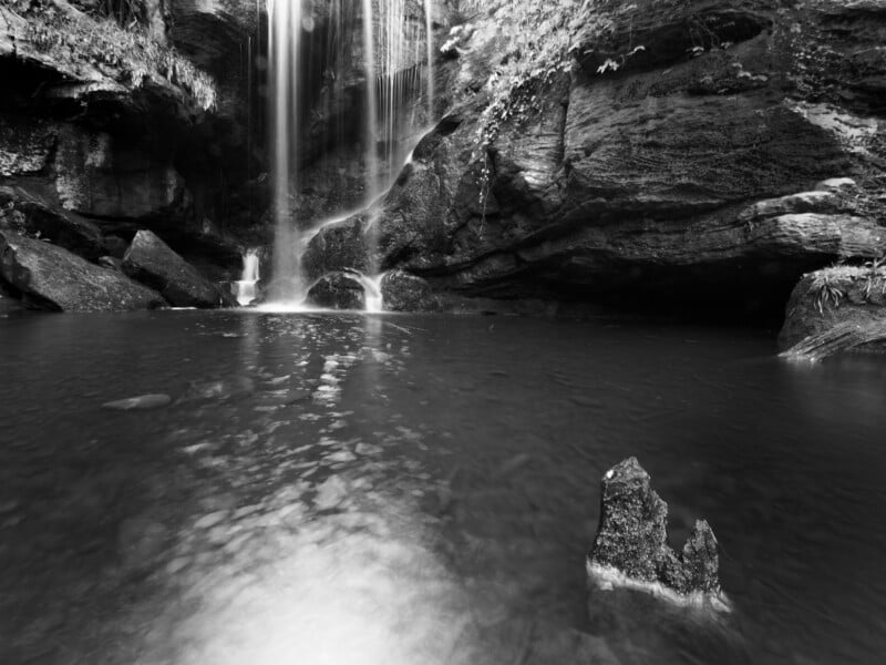 Black and white photo of a waterfall flowing into a calm pool surrounded by rocky cliffs and lush foliage, with a jagged rock protruding from the water in the foreground.