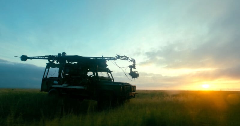 A film camera rig mounted on a vehicle stands in a grassy field at sunset, silhouetted against a dramatic sky with scattered clouds and golden sunlight on the horizon.