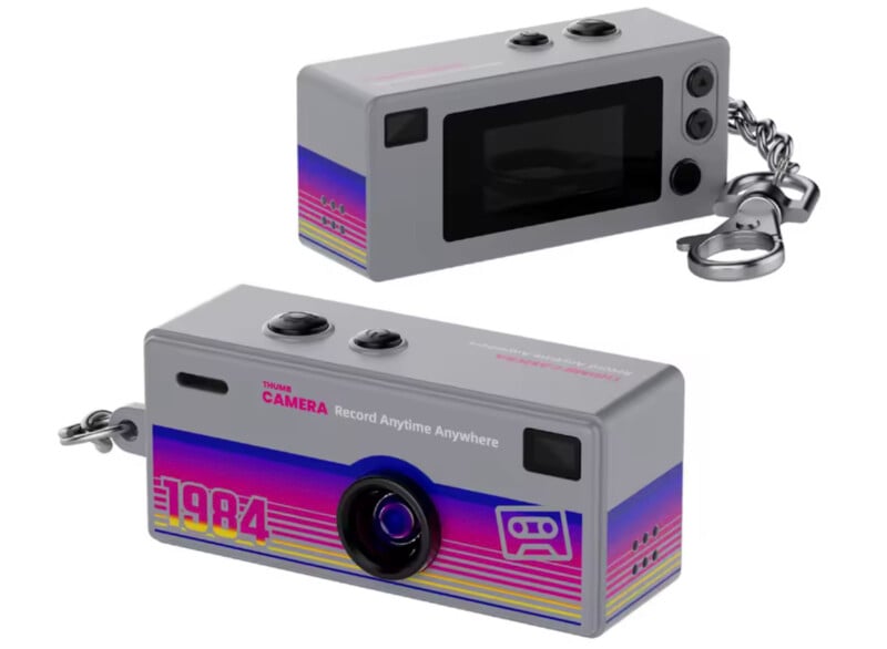 Two views of a small, gray retro-style camera keychain with pink, blue, and yellow stripes. The front has “1984” and cassette graphics; the top shows buttons, and the side displays a keychain attachment.
