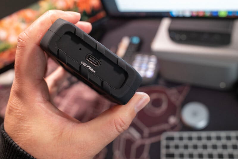 Una mano que sostiene un disco duro externo sólido que muestra un puerto USB 3.0/3.1. El monitor de la computadora, el teclado y otros elementos del escritorio aparecen borrosos en el fondo.