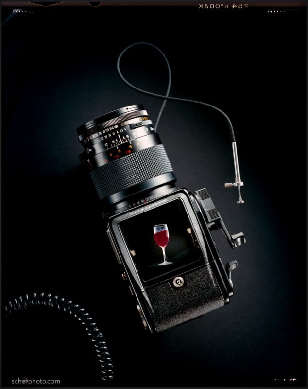 A medium format film camera on a dark surface with its viewfinder showing a clear image of a glass of red wine. A shutter release cable is attached to the camera.