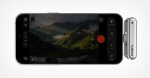 Smartphone in landscape mode displaying a camera app interface, capturing a scenic view of green mountains with sunlight and clouds. A silver device labeled "hohem" is attached to the phone’s side.