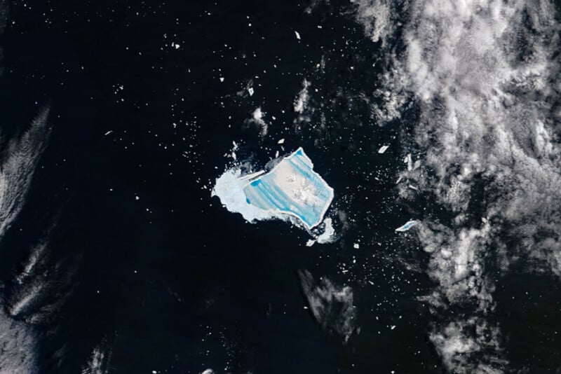 Vista aérea de un enorme iceberg con rayas azules brillantes flotando en aguas oscuras, rodeado de fragmentos de hielo más pequeños y nubes blancas.
