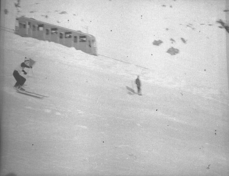 A skier moves downhill on a snowy slope as another person stands in the distance; a train or tram is visible in the background on the snow-covered mountain.