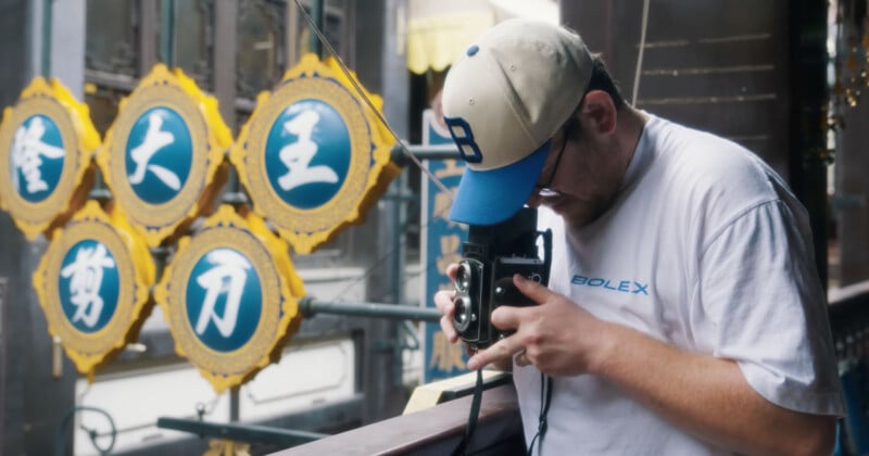 Un hombre con camisa blanca y gorra de béisbol usa una cámara antigua en un balcón, con la ornamentada bandera china amarilla y azul visible al fondo.