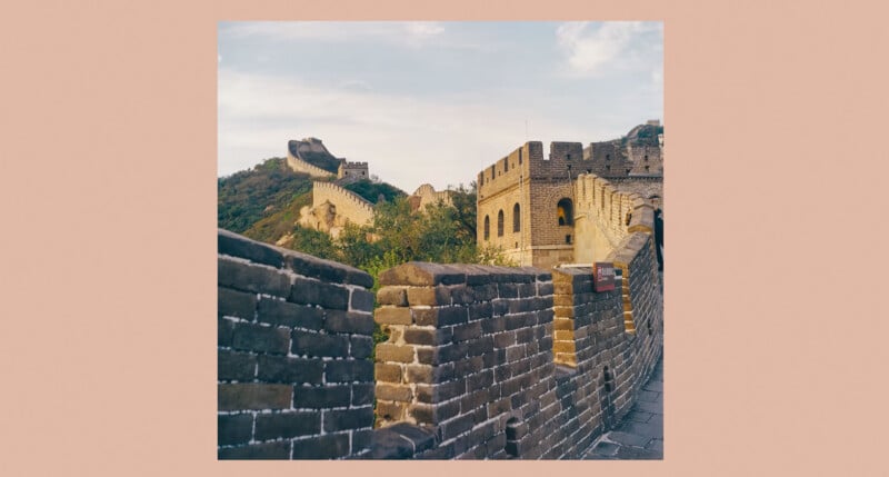 Bajo un cielo parcialmente nublado, una sección de la Gran Muralla China serpentea sobre colinas verdes, con torres de piedra y muros de ladrillo prominentes en primer plano.