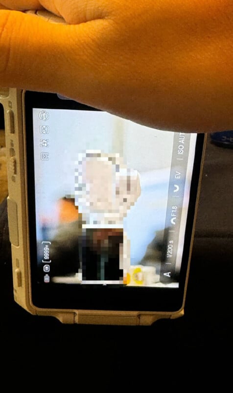 A hand holds a digital camera displaying a pixelated image of a person on the screen, with part of a table and objects visible in the blurred background.
