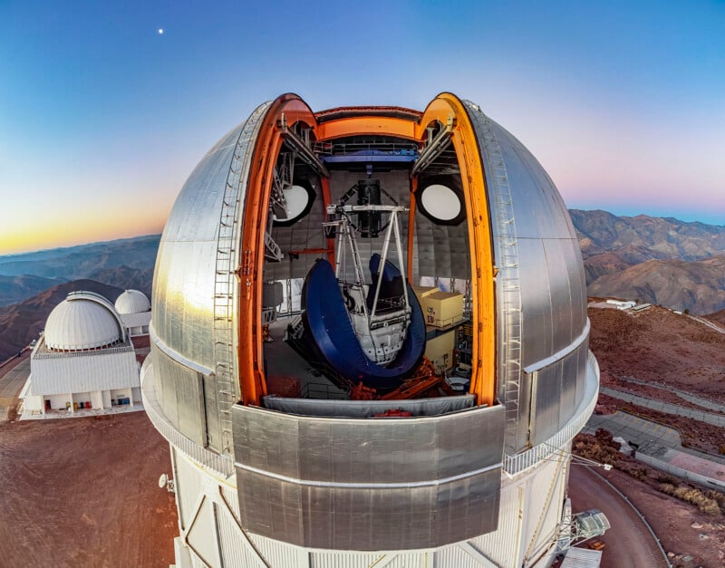 Una enorme cúpula de observatorio con un techo retráctil que se abre para revelar un enorme telescopio en su interior. Al anochecer, aparecen al fondo otras cúpulas del observatorio y un paisaje montañoso escarpado.