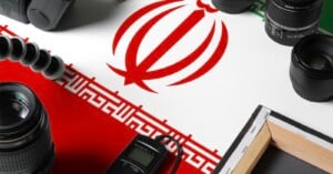 A tabletop with photography equipment, including cameras, lenses, and a tripod, arranged around the flag of Iran, which features a red emblem and green, white, and red stripes.