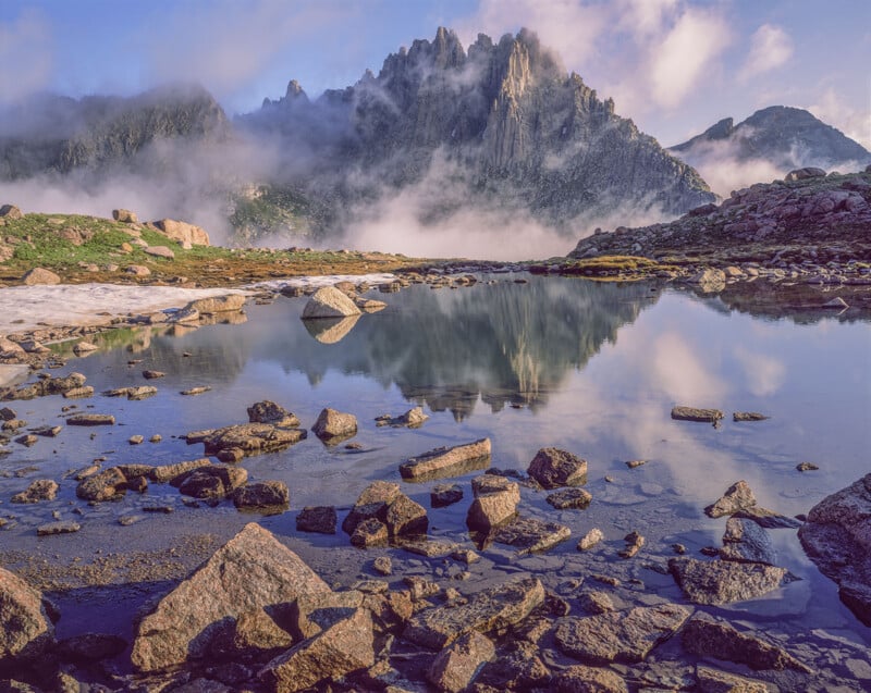 Los picos irregulares que se elevan por encima de las nubes se reflejan en el tranquilo lago de montaña rocosa; piedras y pasto esparcidos rodean el agua, y la niebla y la luz del sol crean una escena tranquila y natural.