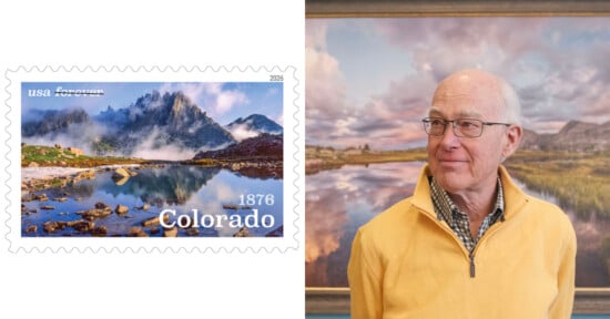 A U.S. postage stamp featuring a scenic Colorado mountain lake is shown next to an older man in glasses and a yellow sweater standing in front of a framed landscape painting.