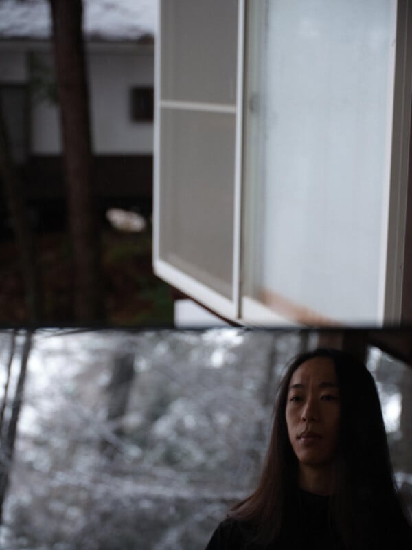 A person with long dark hair stands indoors near a window, looking slightly downward. The background shows a blurry outdoor scene with trees and a building. The atmosphere appears calm and subdued.