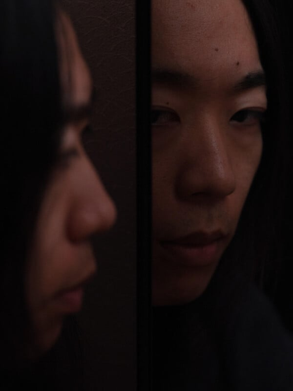 A person with long dark hair looks somberly at their reflection in a mirror, with their face and reflection both visible, creating a moody and introspective atmosphere.