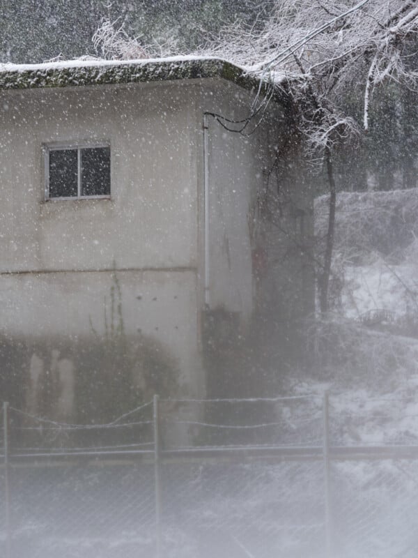 A small building with a single window is partially obscured by heavy snowfall. Snow covers the roof and surrounding ground, while leafless trees and a wire fence are visible in the wintry scene.