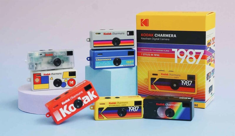 A group of colorful Kodak keychain digital cameras with retro designs are displayed on a pastel surface next to a matching Kodak Charmera 1987 box. The cameras feature various vintage graphics and rainbow stripes.