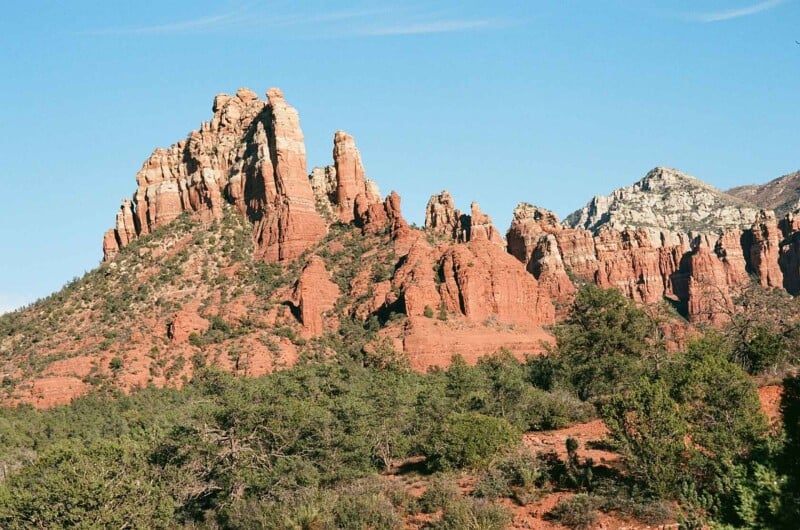 Red rock formations rise sharply from the desert landscape under a clear blue sky, with green shrubs and trees in the foreground. The rocks display layers of red and white, characteristic of a southwestern US desert scene.