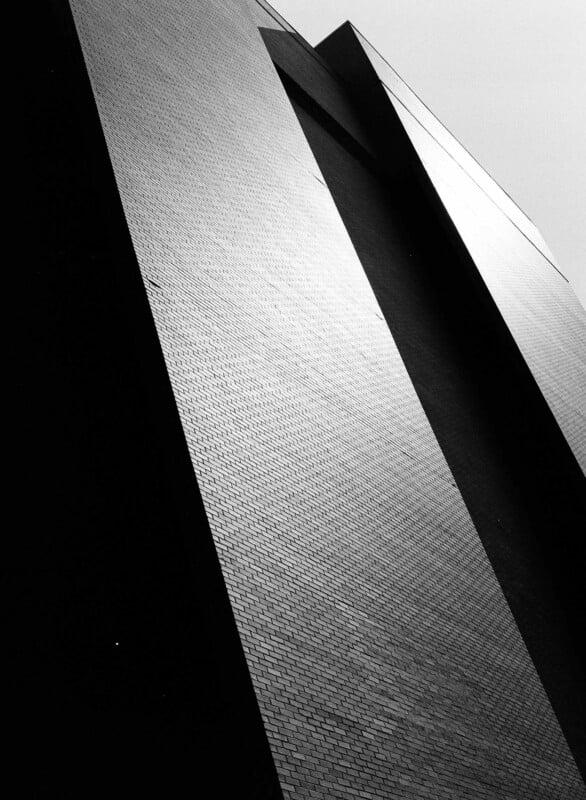 A black and white photo of a tall, modern brick building shot from below, emphasizing sharp angles and geometric patterns, with sunlight casting contrasting shadows on the facade.