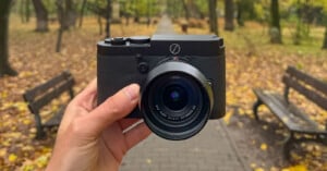 A hand holds a black Leica camera in the center of a park pathway lined with benches and autumn trees, with yellow leaves scattered on the ground.