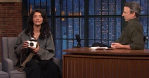 A woman with long dark hair holds a camera while sitting on a talk show set, speaking to a man sitting behind a desk. Cityscape windows and microphones are visible in the background.