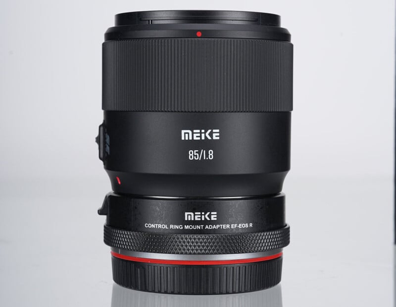 Lente de cámara Meike de 85 mm f/1.8 con adaptador de montura de anillo de control para EF-EOS R, vertical sobre fondo blanco. La lente y el adaptador tienen un cuerpo negro con texto blanco y un anillo decorativo en rojo.