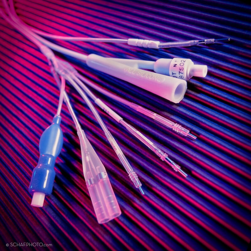 A close-up of various medical catheters and tubes arranged on a ribbed surface, illuminated by pink and blue lighting, highlighting their different shapes and connectors.