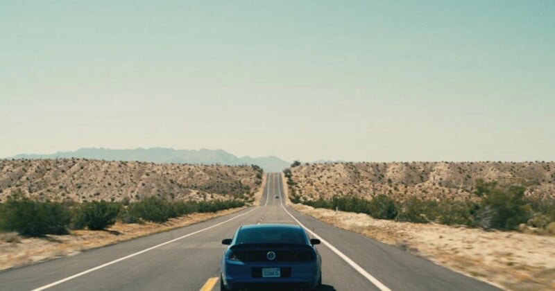 Bajo un cielo despejado, un coche azul circulaba por una carretera recta y vacía del desierto, con montañas distantes claramente visibles en el horizonte. El camino está bordeado de maleza y terreno seco.
