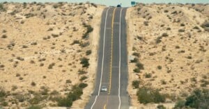 A long, straight road stretches through a dry, sandy desert landscape with sparse shrubs. One white car drives towards the horizon, while another vehicle is barely visible at the top of a hill ahead.