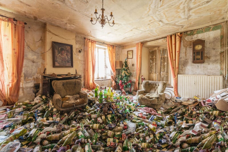 A cluttered, abandoned living room with worn armchairs, orange curtains, and a chandelier. The floor is covered with piles of empty bottles and trash, creating a chaotic scene. Light enters through two windows.
