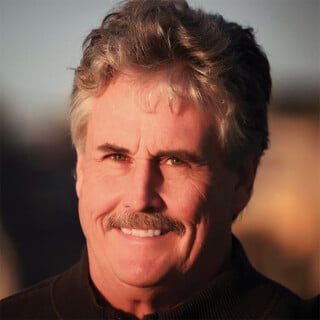 A middle-aged man with wavy gray hair and a mustache smiles at the camera. He is wearing a dark jacket, and the background is softly blurred with warm lighting.