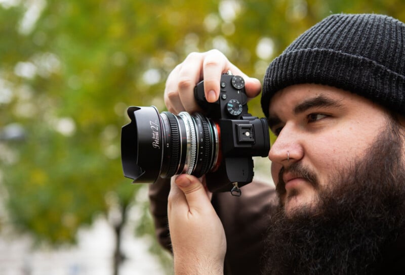 A bearded person wearing a black beanie is holding a camera up to their face, taking a photo outdoors with blurred green foliage in the background.