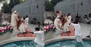A wedding photoshoot by a pool. A photographer accidentally falls into the water as the bride and groom, dressed in traditional attire, pose with two men on a colorful rug near a flower arrangement.