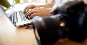 Close-up of hands typing on a laptop keyboard with a professional camera and a memory card on the wooden table in the foreground.
