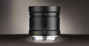 A black camera lens with engraved focus and aperture markings, including a yellow "75" for the focal length, sits upright against a gradient dark background. The lens features a textured grip and metallic mounting ring.