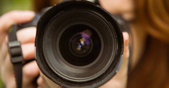 Close-up of a person holding a camera, focusing on the camera lens. The face is out of focus in the background, emphasizing the details of the lens and the act of taking a photo.
