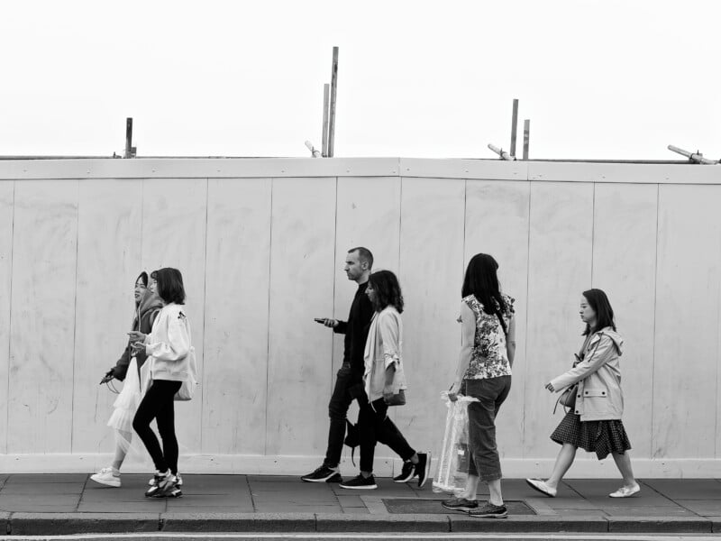 A black and white photo of six people walking on a sidewalk in front of a construction barrier, all moving in the same direction; some are carrying bags and looking ahead.