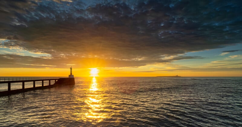 El amanecer en el mar, la luz dorada se refleja en el agua, hay un pequeño faro en el muelle de la izquierda y el cielo está lleno de nubes espectaculares.