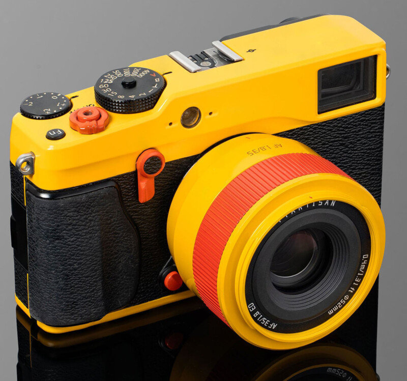 Cámara digital de estilo retro con cuerpo amarillo brillante, mango texturizado negro, lente amarilla, anillo de enfoque rojo, colocada sobre una superficie reflectante negra.