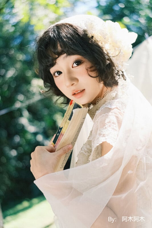 Una mujer joven con el pelo corto y oscuro, vestida con un vestido de encaje blanco y un tocado floral, sostiene un libro y mira a la cámara. La luz del sol brilla a través de las hojas verdes del fondo.