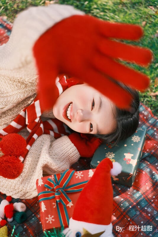 Una mujer sonriente con un suéter acogedor y guantes rojos camina hacia la cámara rodeada de regalos de Navidad envueltos en una manta festiva.