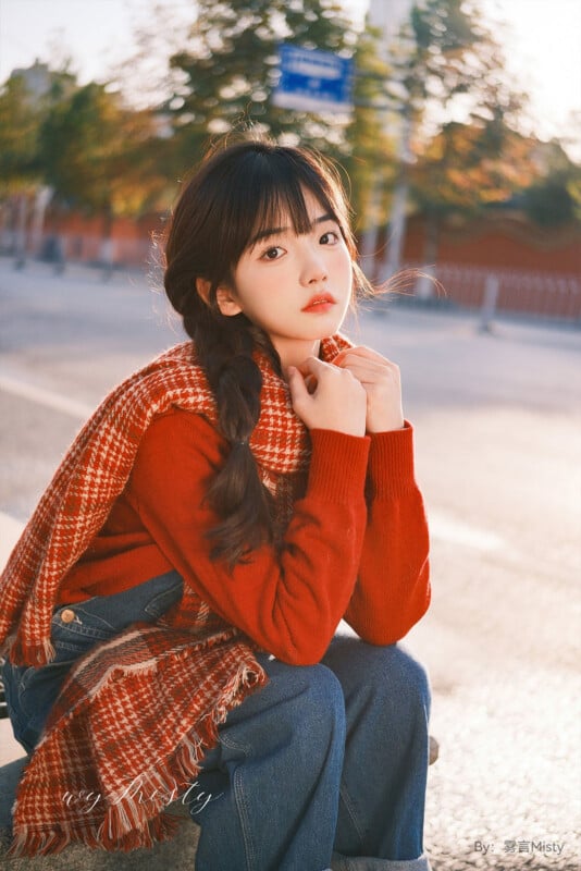 En un día soleado, una mujer joven con el pelo largo y negro trenzado, vestida con un suéter rojo y una bufanda a cuadros estaba sentada al aire libre, apoyando la barbilla en la mano y mirando pensativamente a la cámara. El fondo son árboles y una pared roja.