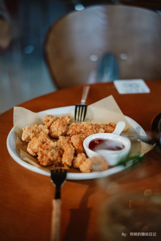 Un plato de nuggets de pollo frito sobre papel pergamino con un plato pequeño de salsa roja sobre una mesa de madera con un tenedor y un fondo borroso.