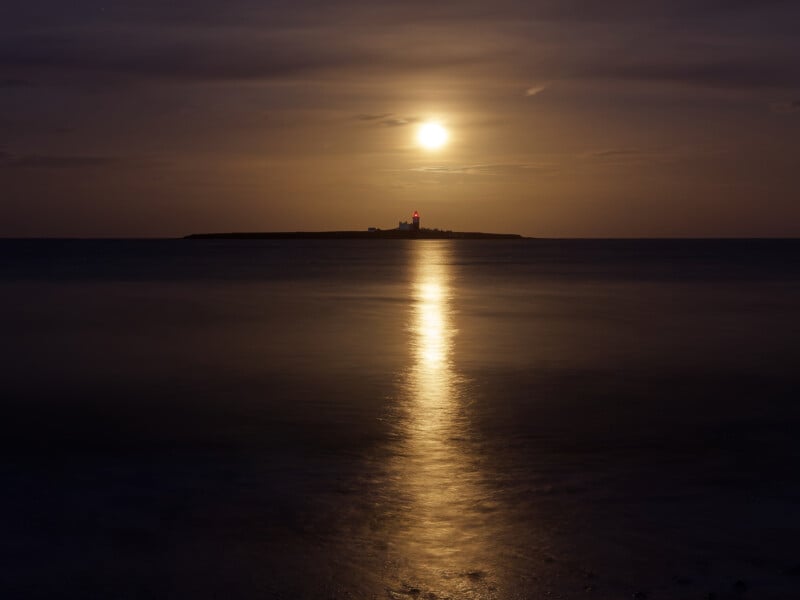 A lighthouse on a distant island is silhouetted against a golden sky, with the moon shining above and its reflection stretching across calm, dark water at night.