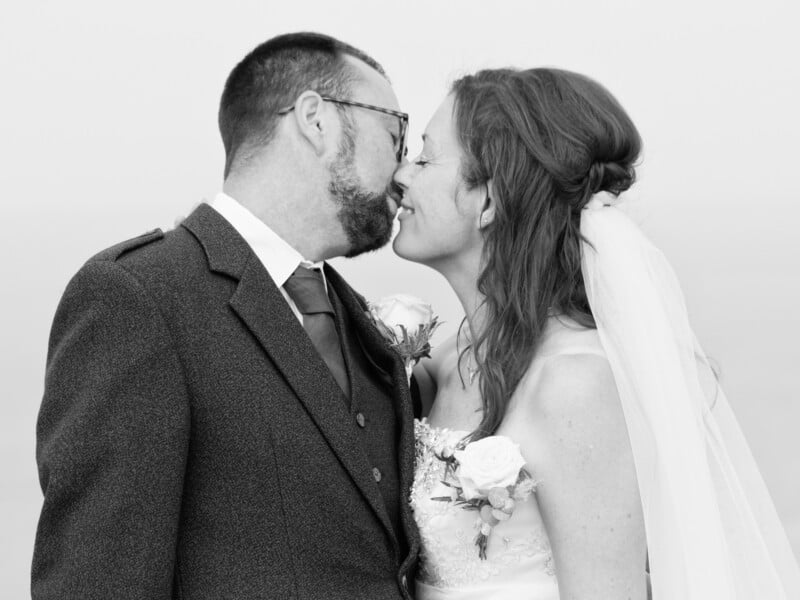A man in a suit and a woman in a wedding dress with a veil smile and touch noses lovingly, holding each other close. Both wear boutonnieres, and the photo is in black and white.