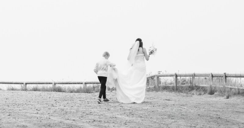 A bride in a wedding dress walks outdoors on a dirt path, holding a bouquet. A young boy walks beside her, holding part of her dress. A wooden fence lines the background. The image is in black and white.